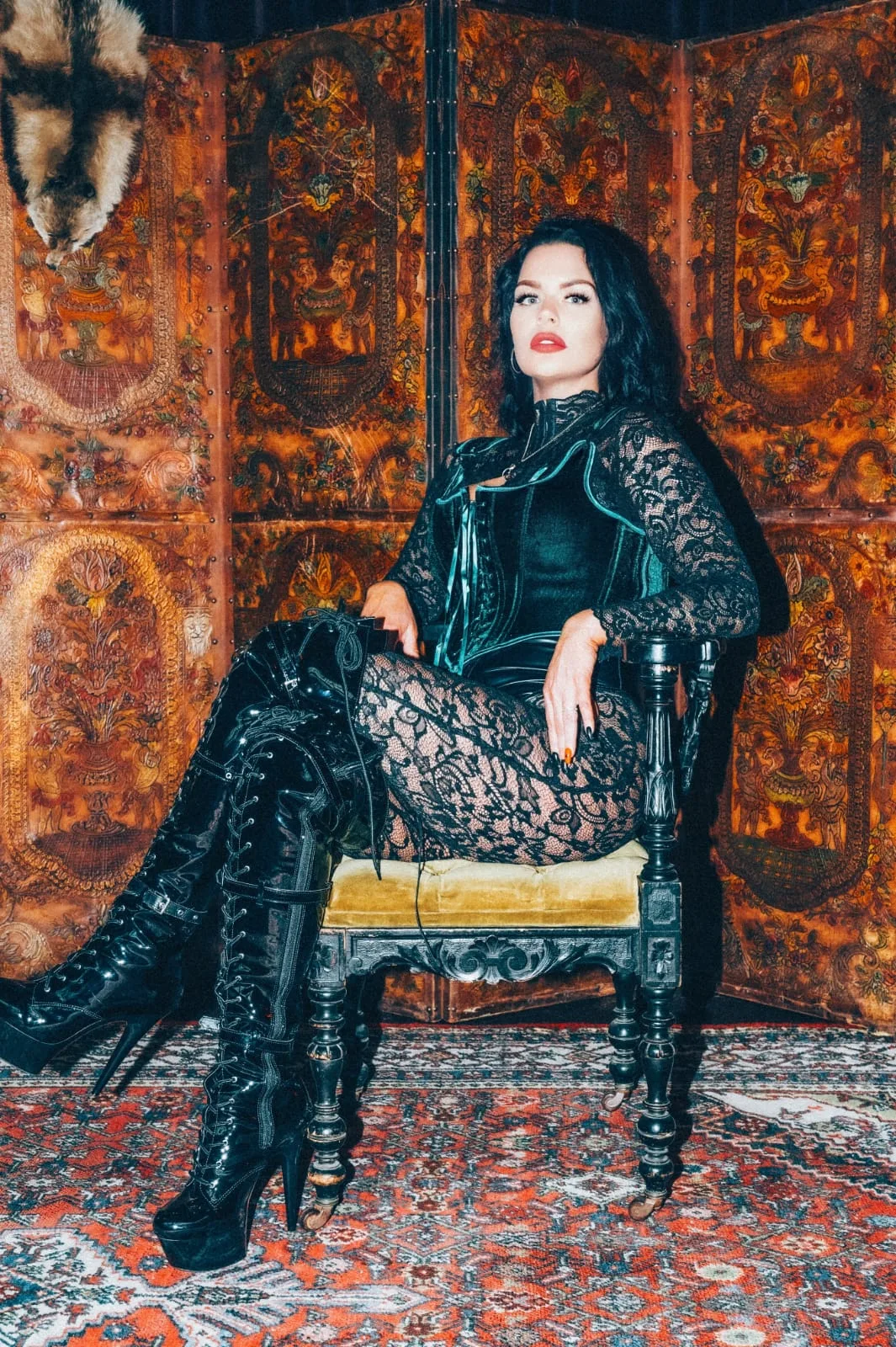 Mistress Charlotte O’Winter, professional dominatrix in London, seated in black lace and leather boots before a carved screen