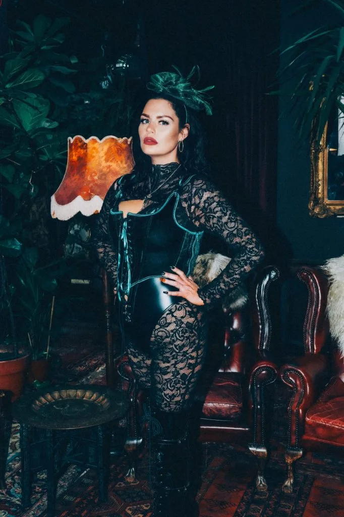 Mistress Charlotte standing beside an amber lamp in a leather corset and black lace