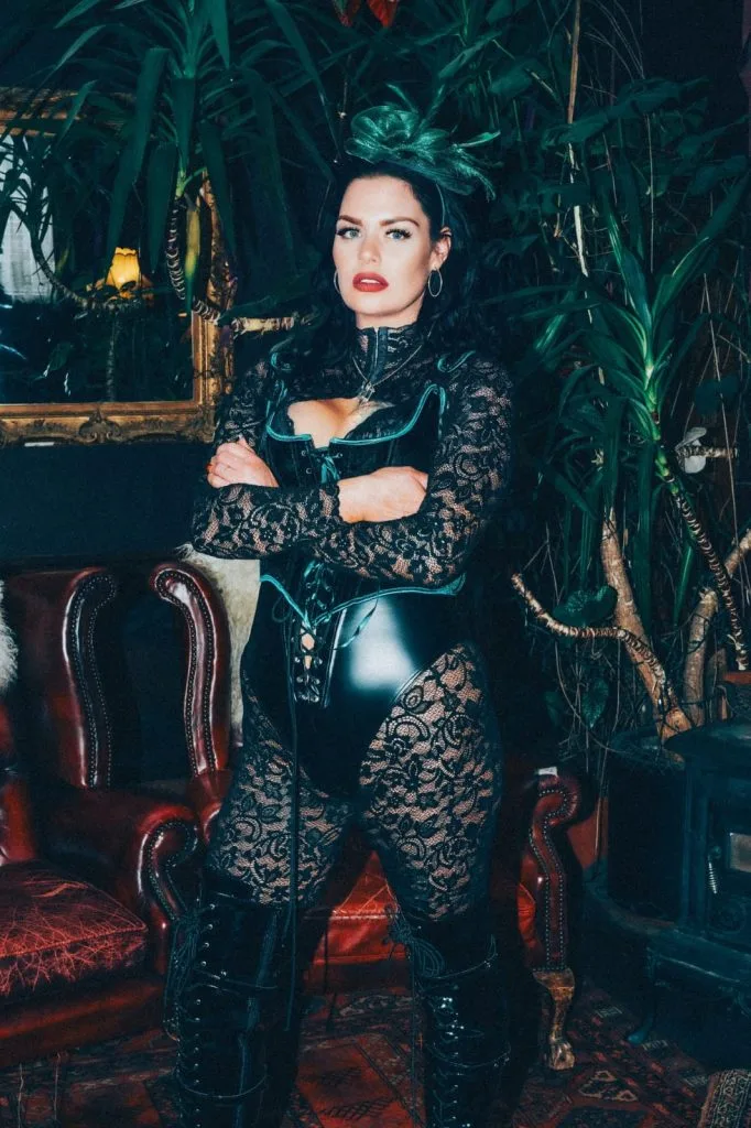 Mistress Charlotte with arms folded among palms, in a black corset and lace