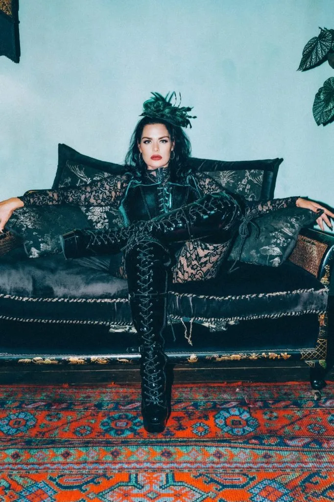 Mistress Charlotte seated with her arms outstretched along a black sofa, wearing a feathered headpiece