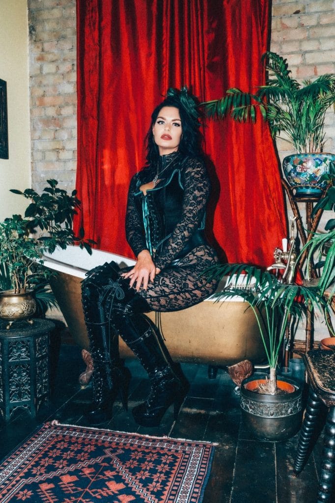 Mistress Charlotte O’Winter seated before a red velvet curtain in black lace, corset and thigh-high boots