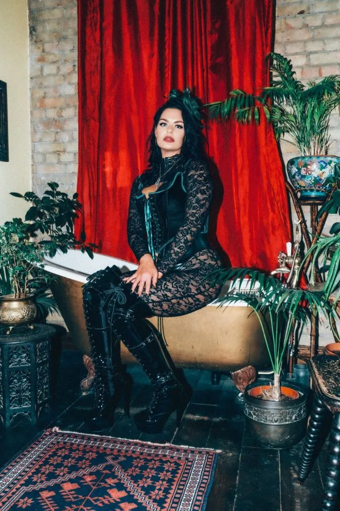 Mistress Charlotte seated on the edge of a bath before a red velvet curtain