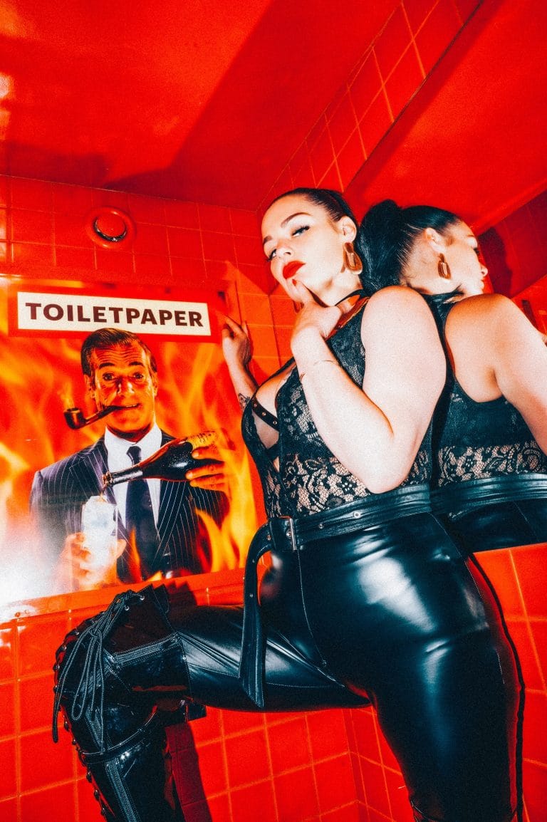 Two dominatrices in black lace and latex posing against a red tiled wall